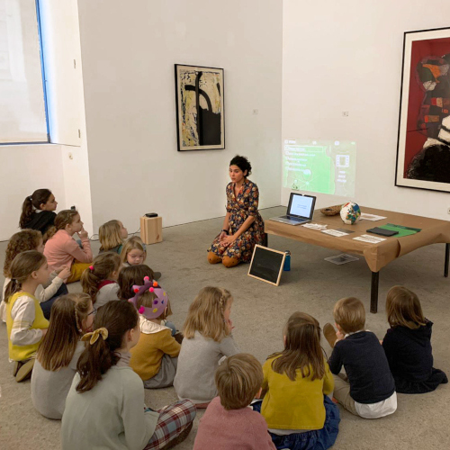 Vista lateral de María en un taller frente a un grupo de niños en una galería de arte.